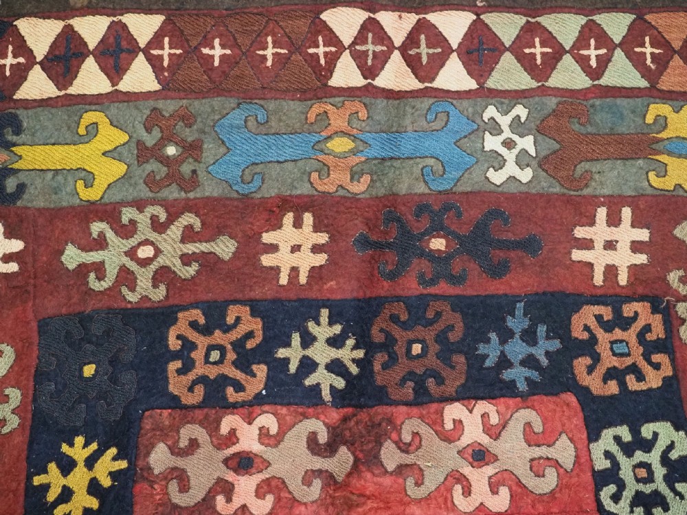 An antique Central Asian embroidered felt rug featuring a terracotta red centre with diamond medallions surrounded by multi-coloured geometric borders on dark brown felt. View X