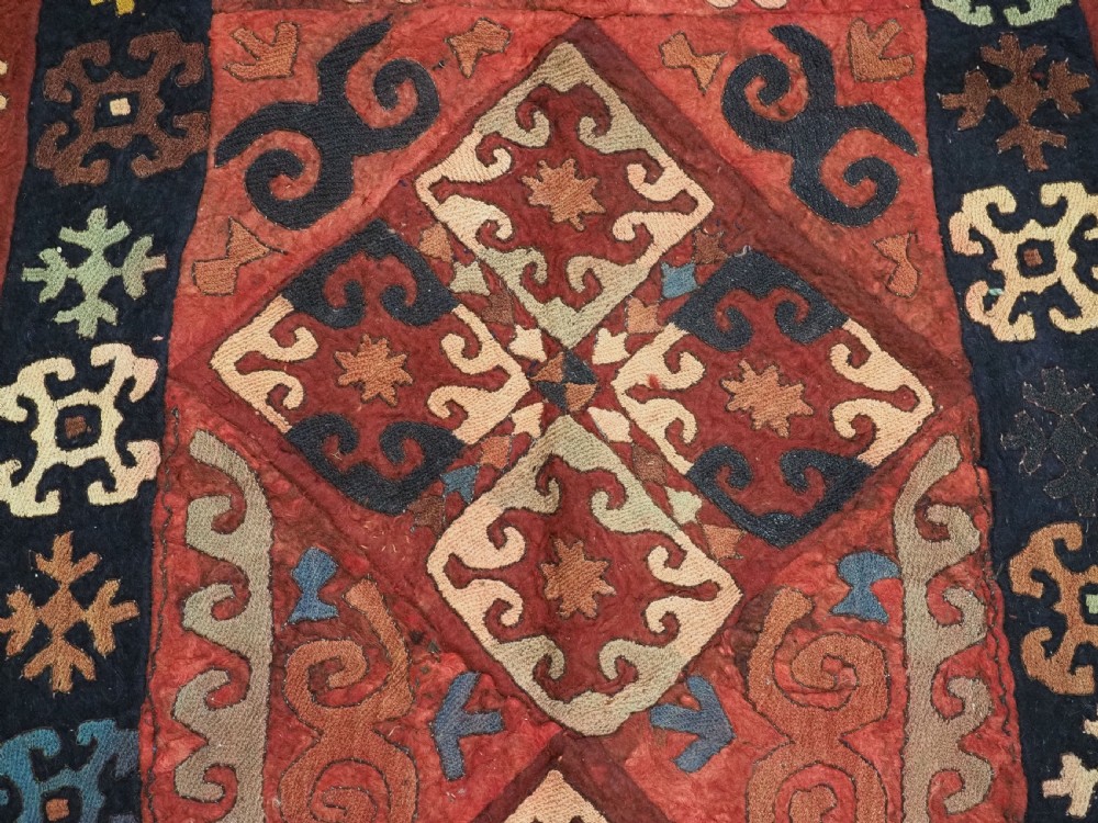 An antique Central Asian embroidered felt rug featuring a terracotta red centre with diamond medallions surrounded by multi-coloured geometric borders on dark brown felt. View XI