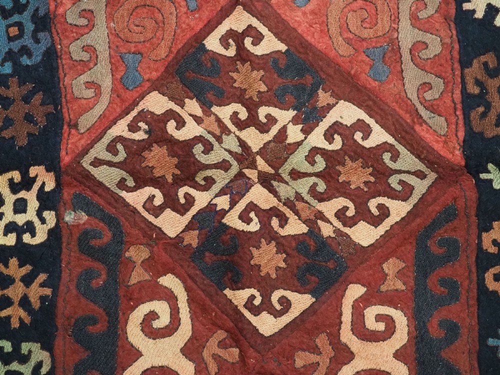 An antique Central Asian embroidered felt rug featuring a terracotta red centre with diamond medallions surrounded by multi-coloured geometric borders on dark brown felt. View XII