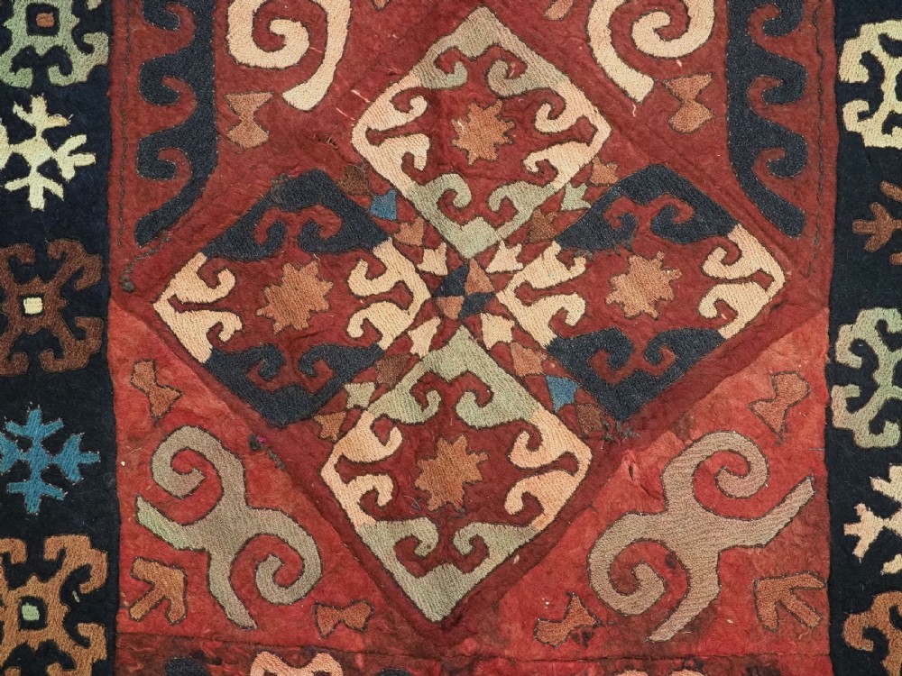 An antique Central Asian embroidered felt rug featuring a terracotta red centre with diamond medallions surrounded by multi-coloured geometric borders on dark brown felt. View XIII