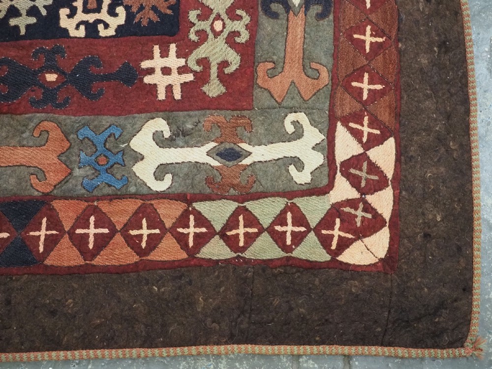 An antique Central Asian embroidered felt rug featuring a terracotta red centre with diamond medallions surrounded by multi-coloured geometric borders on dark brown felt. View XV