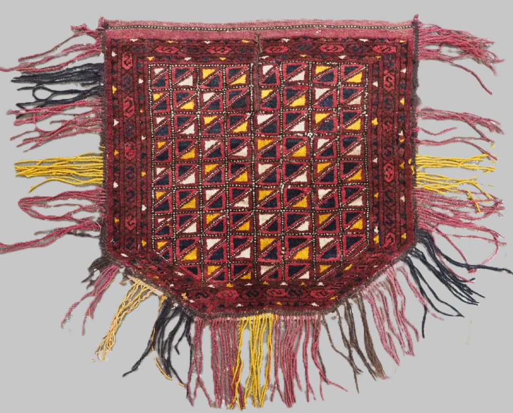 An antique Ersari Turkmen saddle cover rug with a grid pattern of triangular motifs in red, navy, yellow, and white, trimmed with multi-coloured yarn tassels.