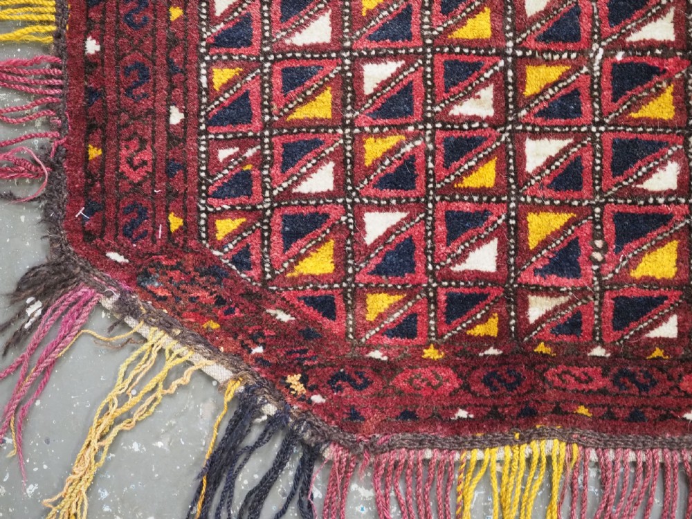An antique Ersari Turkmen saddle cover rug with a grid pattern of triangular motifs in red, navy, yellow, and white, trimmed with multi-coloured yarn tassels. View IV