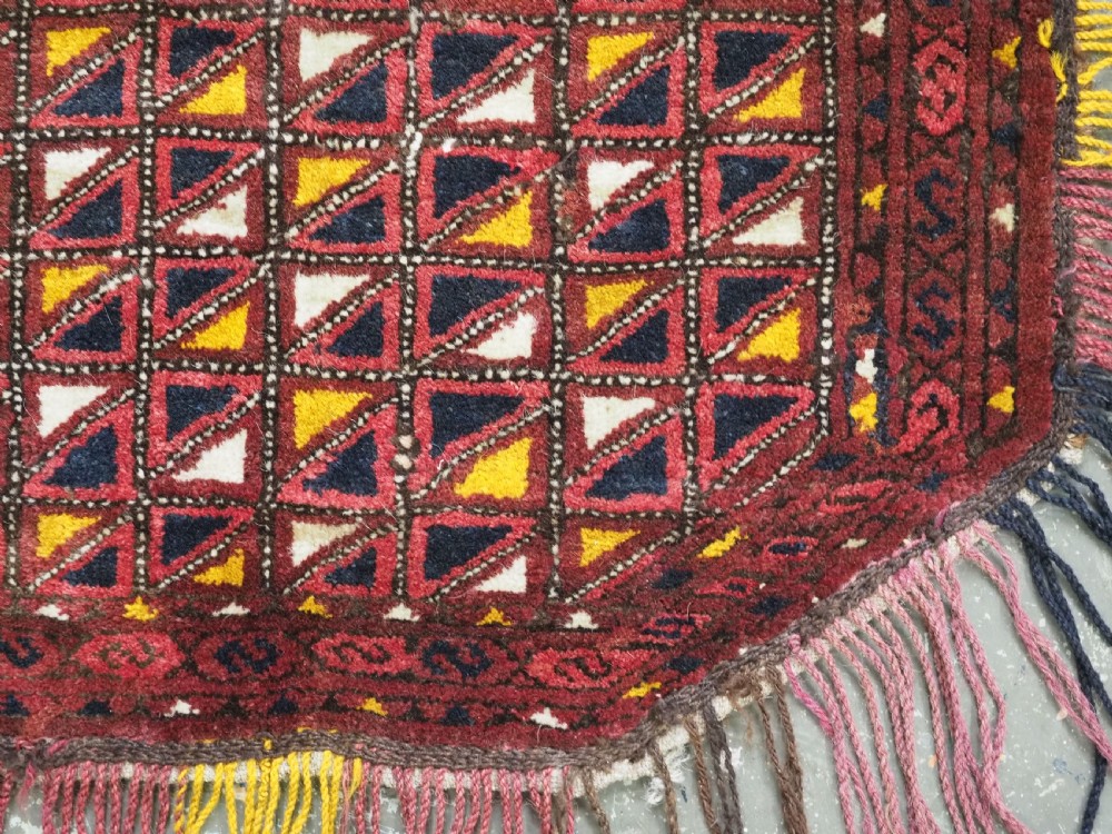 An antique Ersari Turkmen saddle cover rug with a grid pattern of triangular motifs in red, navy, yellow, and white, trimmed with multi-coloured yarn tassels. View V