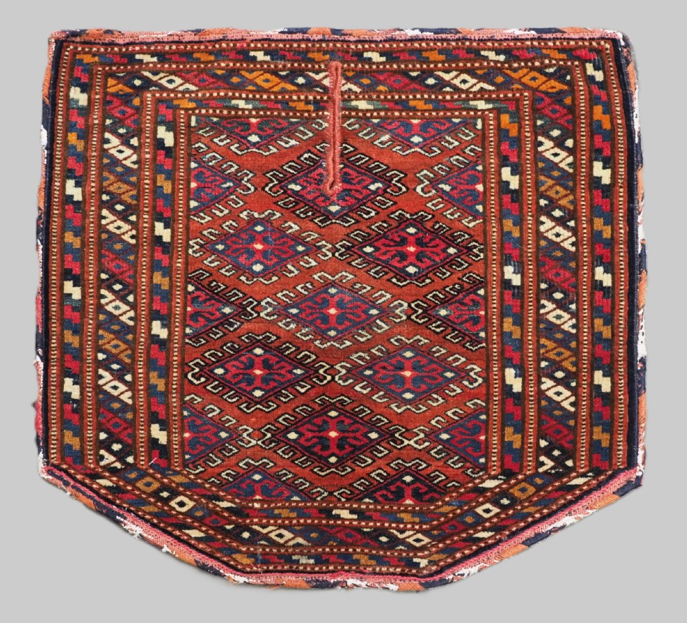 A handwoven Yomut Turkmen wool saddle cover featuring geometric diamond motifs in terracotta red, dark blue, and ivory with a central saddle horn slit.