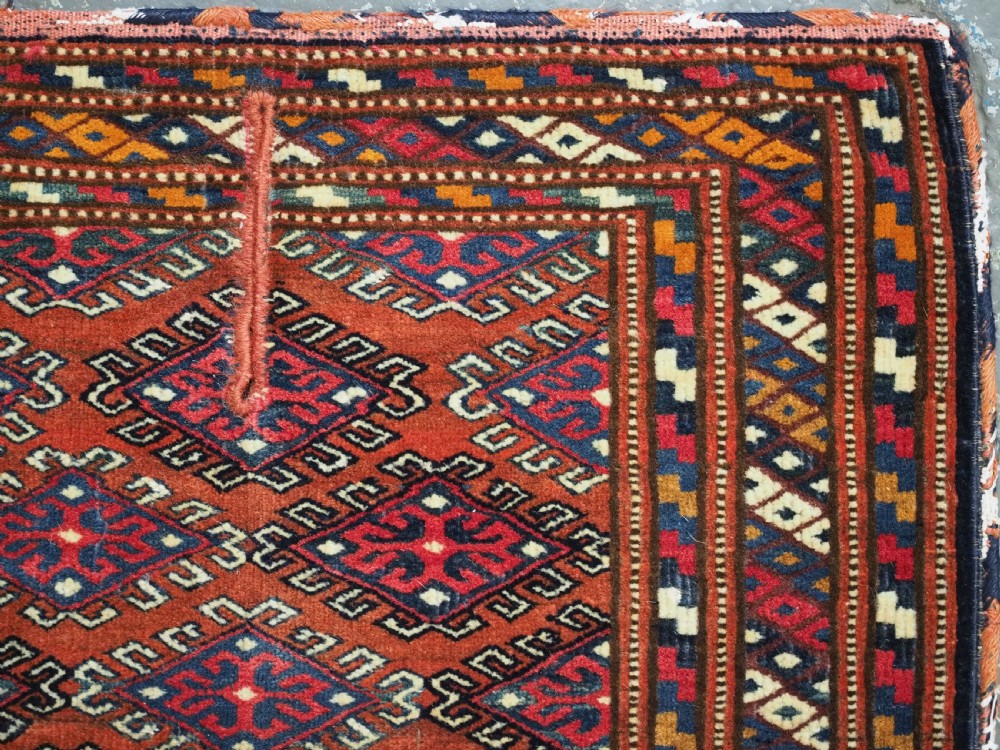 A handwoven Yomut Turkmen wool saddle cover featuring geometric diamond motifs in terracotta red, dark blue, and ivory with a central saddle horn slit. View III