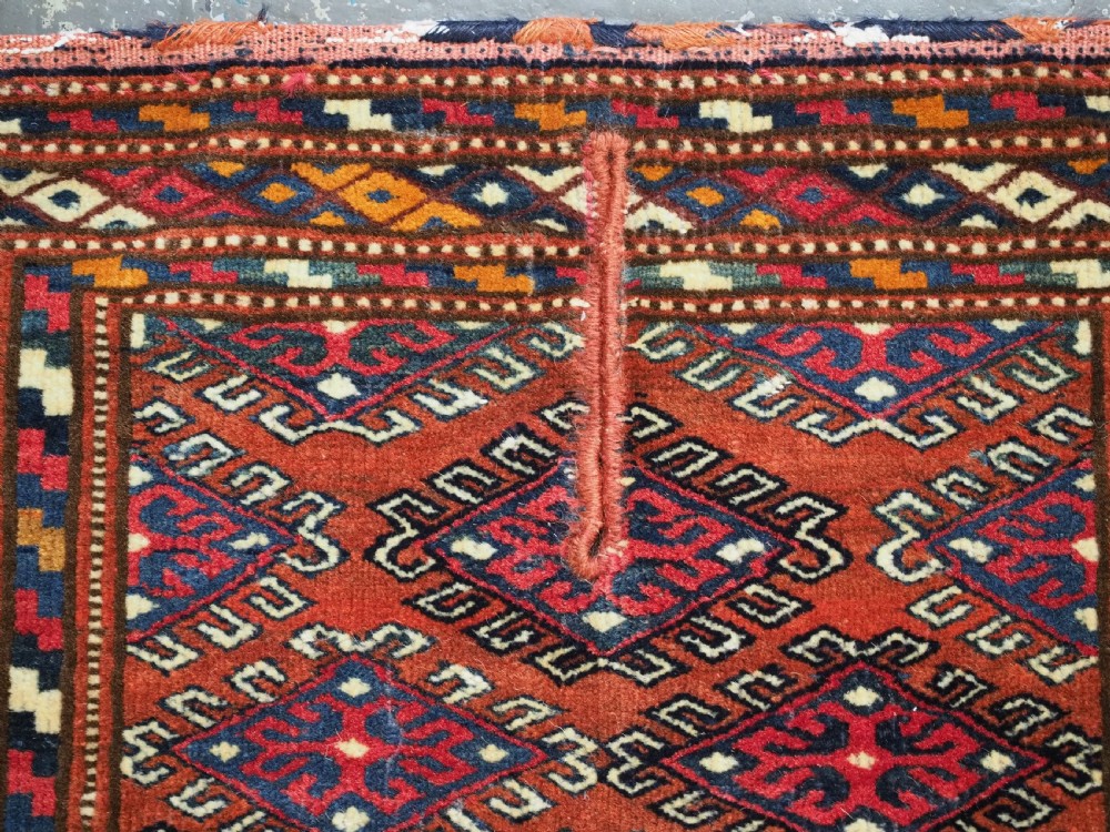 A handwoven Yomut Turkmen wool saddle cover featuring geometric diamond motifs in terracotta red, dark blue, and ivory with a central saddle horn slit. View VI