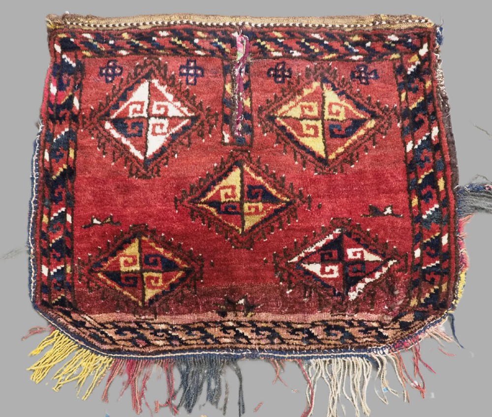 A square antique woven wool saddle cover featuring a crimson field decorated with five geometric diamond motifs and multi-coloured fringe along the bottom edge.