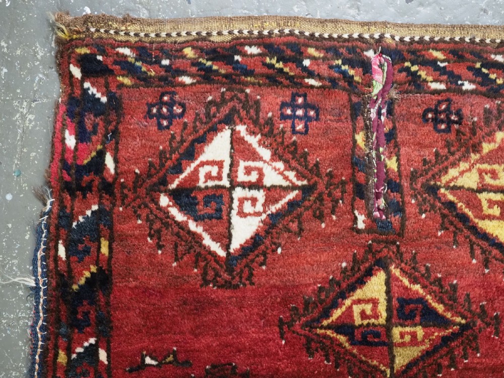 A square antique woven wool saddle cover featuring a crimson field decorated with five geometric diamond motifs and multi-coloured fringe along the bottom edge. View II