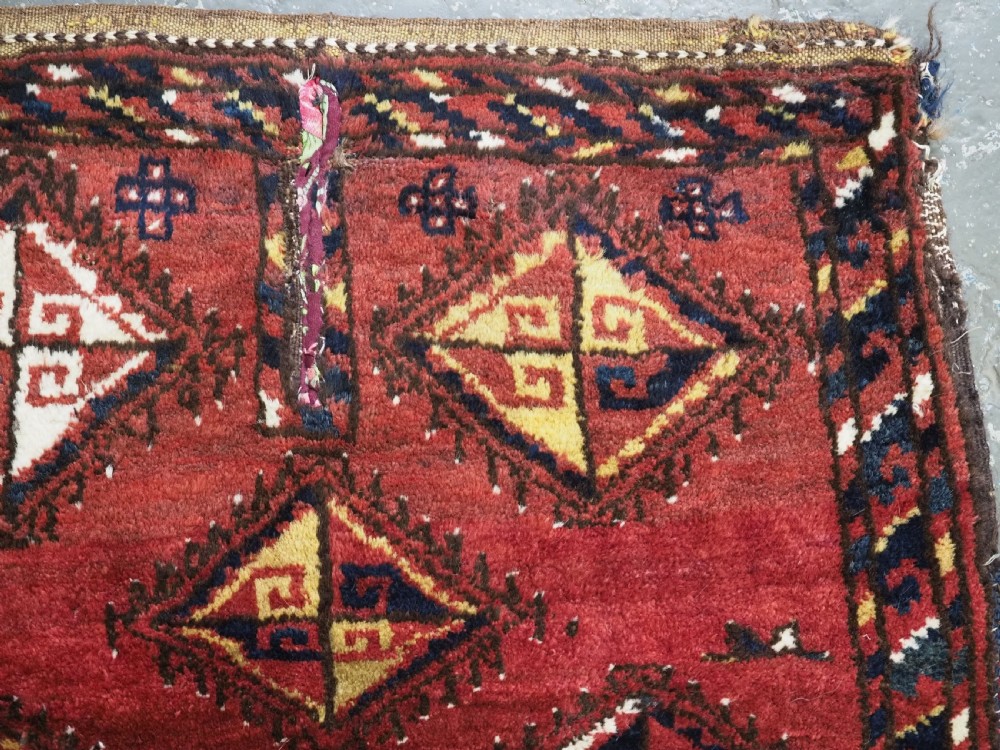 A square antique woven wool saddle cover featuring a crimson field decorated with five geometric diamond motifs and multi-coloured fringe along the bottom edge. View III