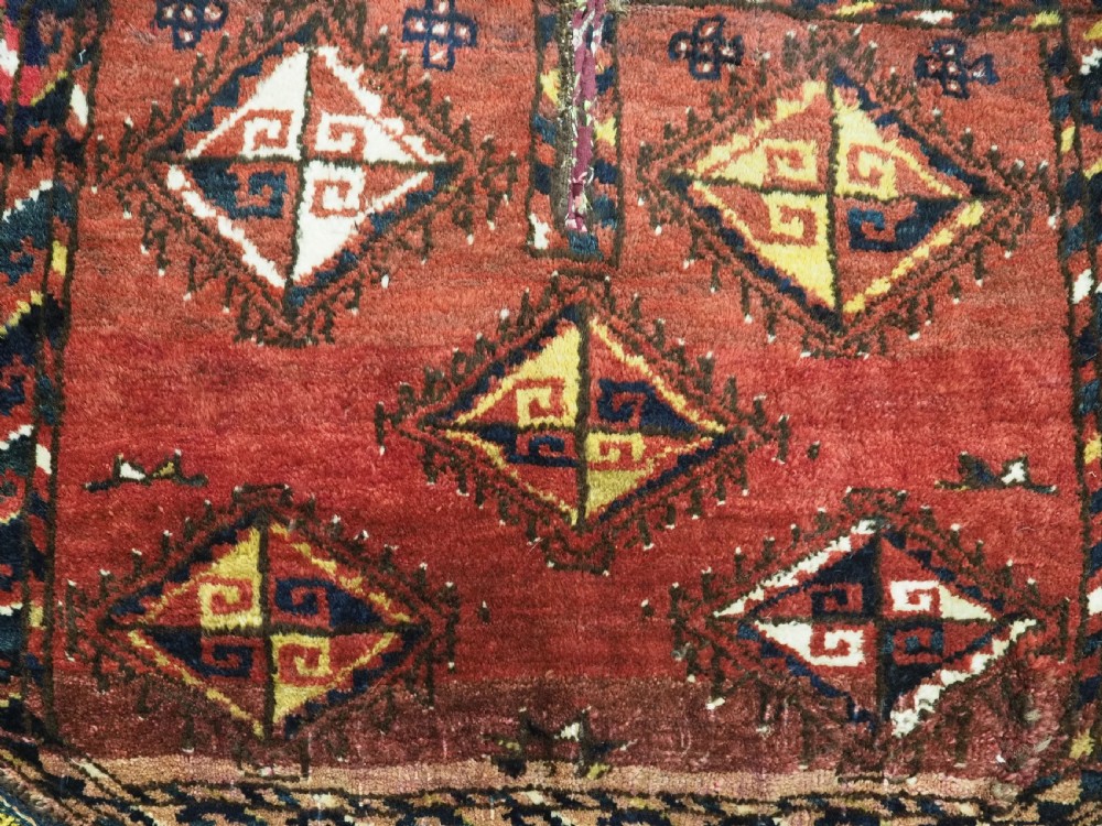 A square antique woven wool saddle cover featuring a crimson field decorated with five geometric diamond motifs and multi-coloured fringe along the bottom edge. View IV