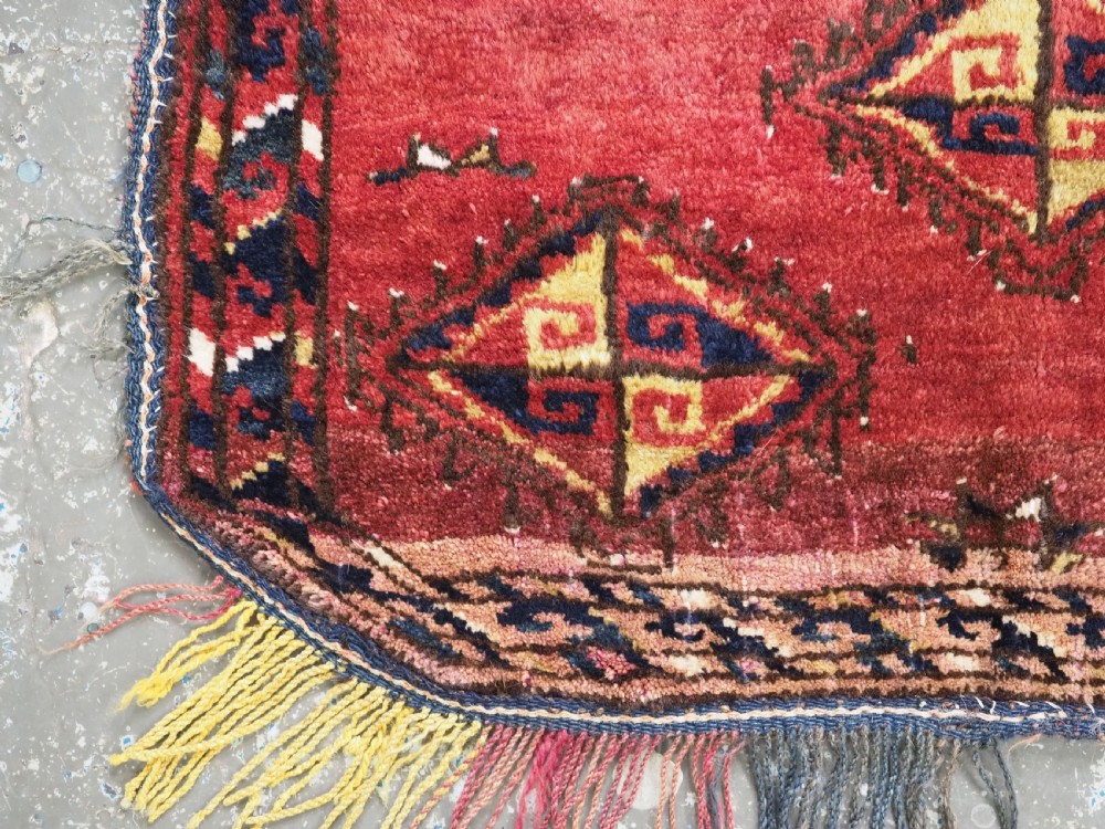 A square antique woven wool saddle cover featuring a crimson field decorated with five geometric diamond motifs and multi-coloured fringe along the bottom edge. View V
