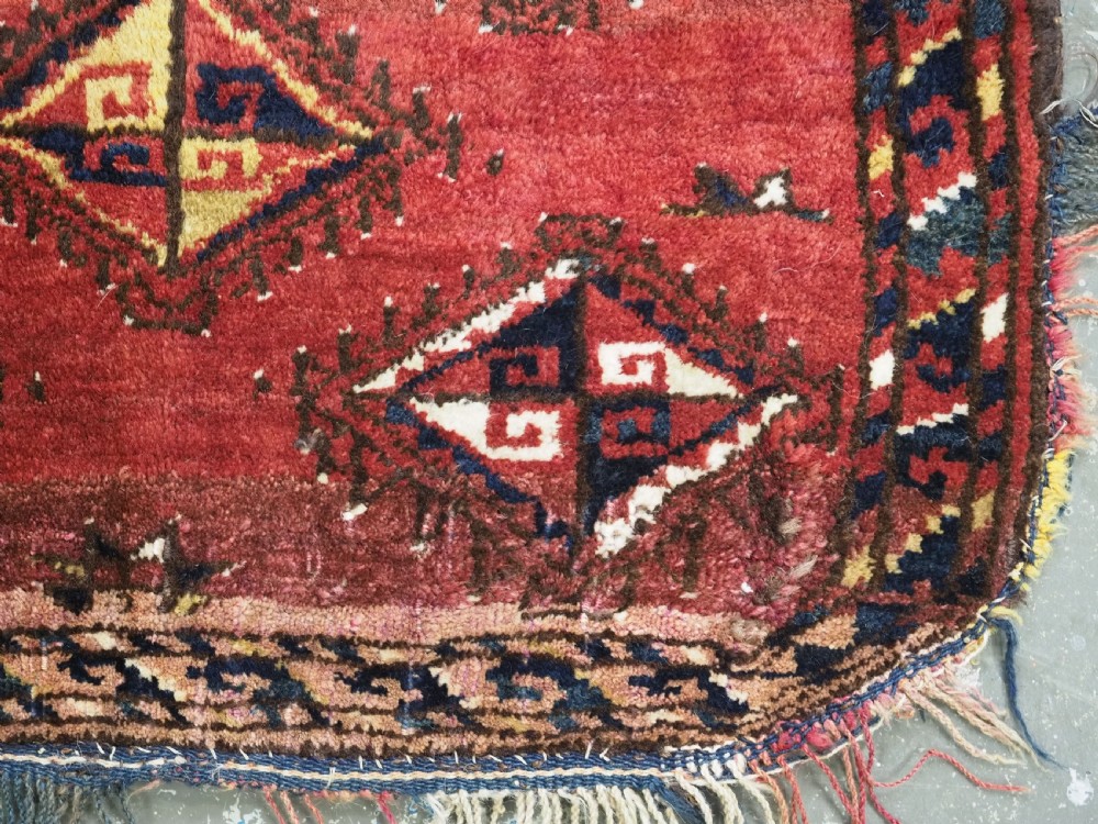 A square antique woven wool saddle cover featuring a crimson field decorated with five geometric diamond motifs and multi-coloured fringe along the bottom edge. View VI