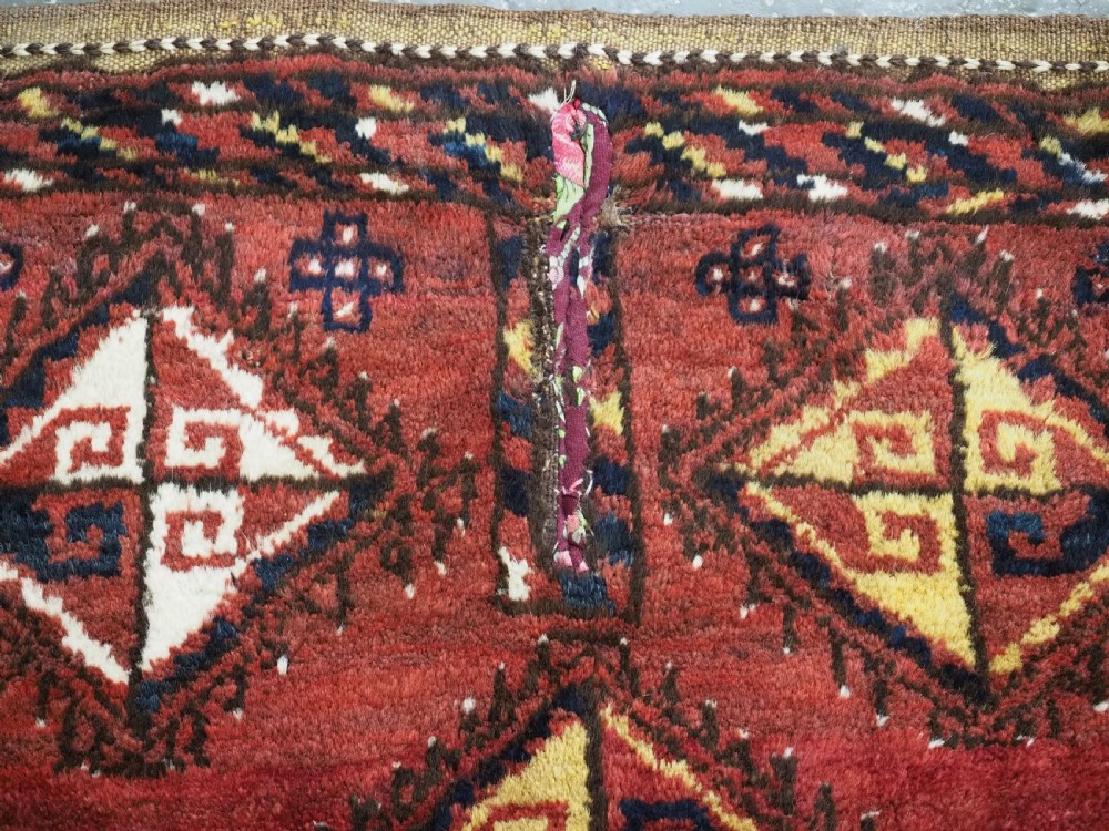 A square antique woven wool saddle cover featuring a crimson field decorated with five geometric diamond motifs and multi-coloured fringe along the bottom edge. View VII