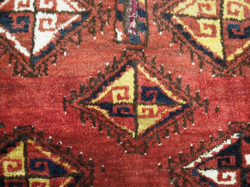 A square antique woven wool saddle cover featuring a crimson field decorated with five geometric diamond motifs and multi-coloured fringe along the bottom edge. View VIII