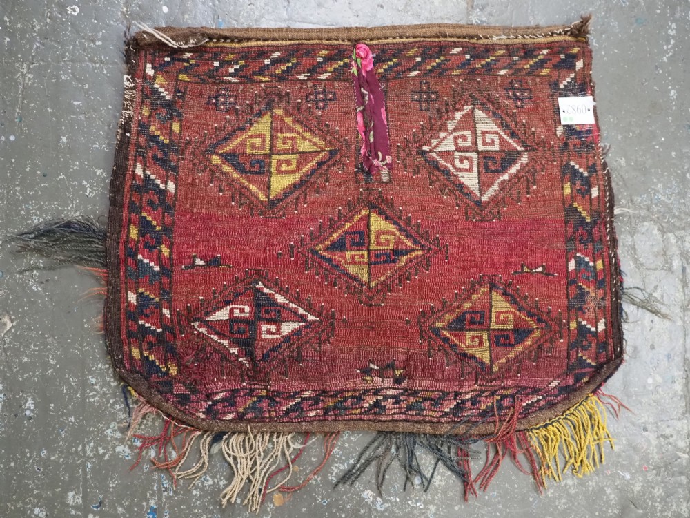 A square antique woven wool saddle cover featuring a crimson field decorated with five geometric diamond motifs and multi-coloured fringe along the bottom edge. View IX