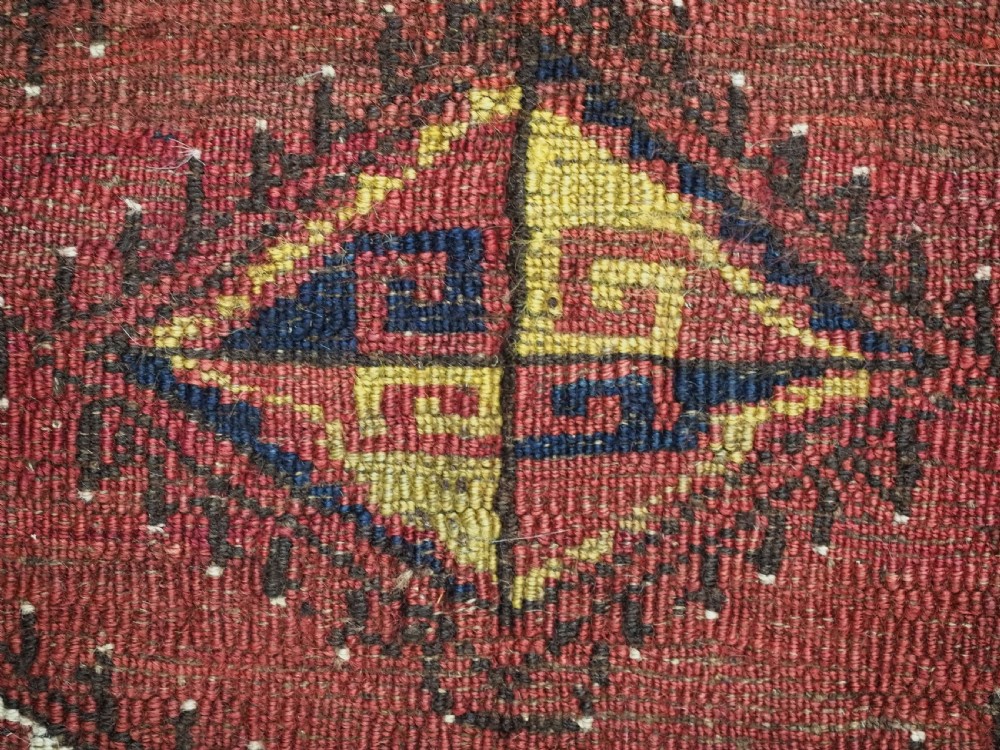 A square antique woven wool saddle cover featuring a crimson field decorated with five geometric diamond motifs and multi-coloured fringe along the bottom edge. View X