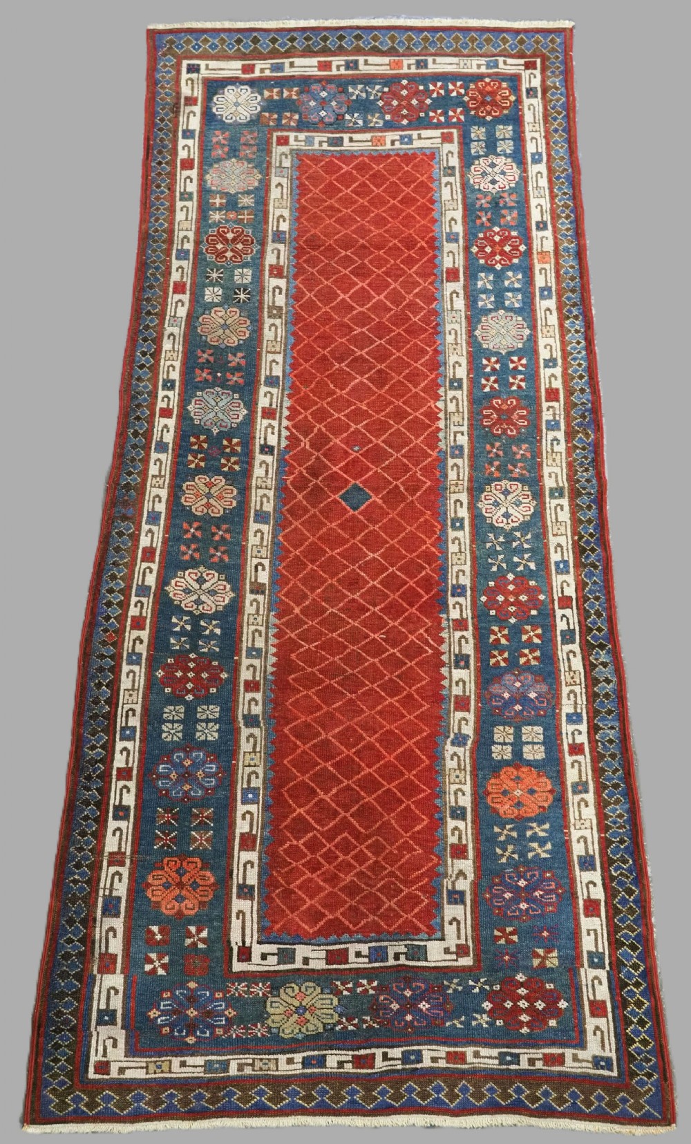 An antique Caucasian Talish runner featuring a terracotta red lattice field surrounded by a wide indigo blue border filled with geometric rosettes.