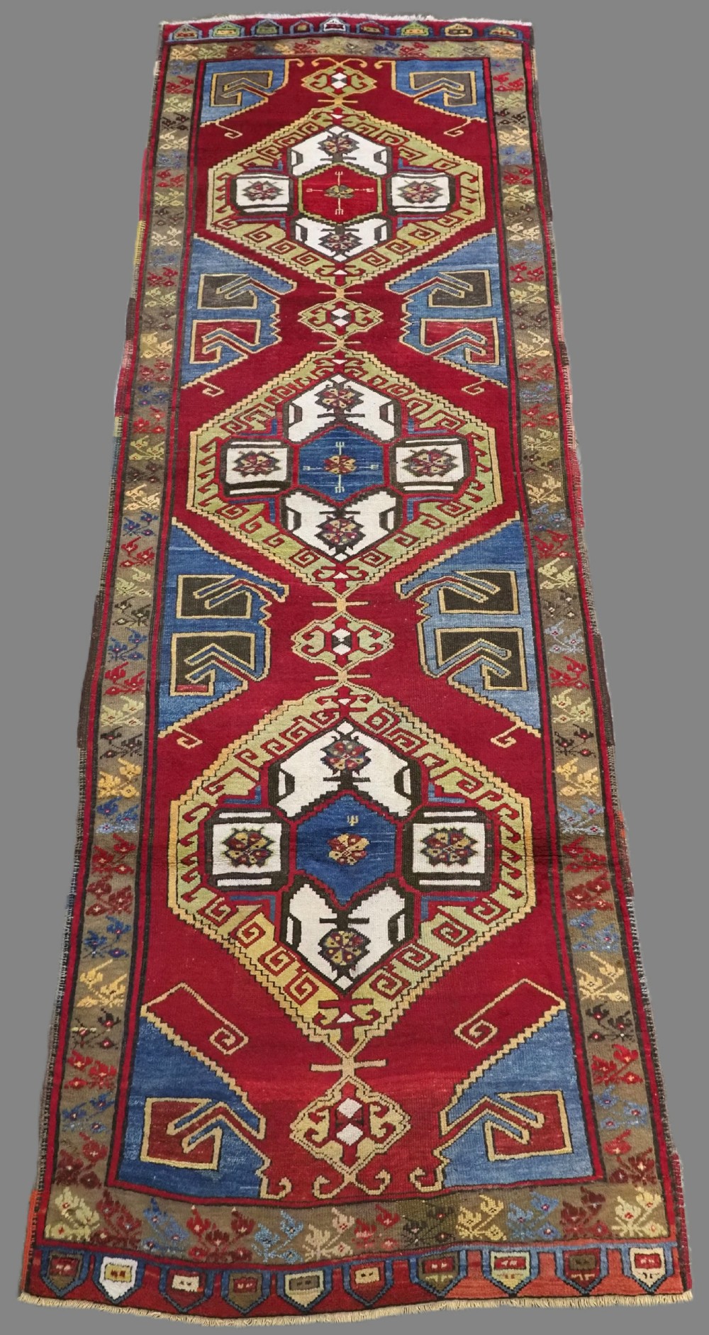 An antique Turkish long wool rug featuring three primary geometric medallions on a vibrant red field with blue and patterned border panels.