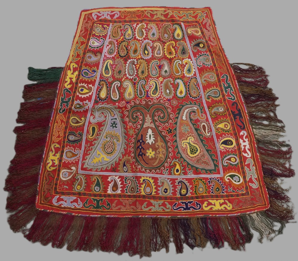 An antique trapezoidal red textile cover featuring dense paisley chain-stitch embroidery in yellow, green, and cream, finished with heavy fringe along the edges.