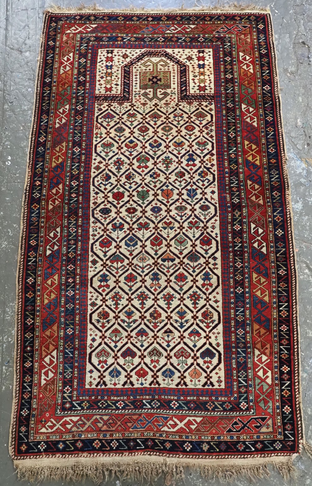 An antique Caucasian Dagestan prayer rug featuring an ivory field with a floral lattice pattern, a central mihrab arch, and multiple geometric borders in red and navy blue.