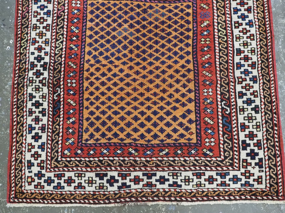 A small square antique hand-knotted wool rug featuring a mustard yellow lattice central field surrounded by red and ivory geometric borders. View IV
