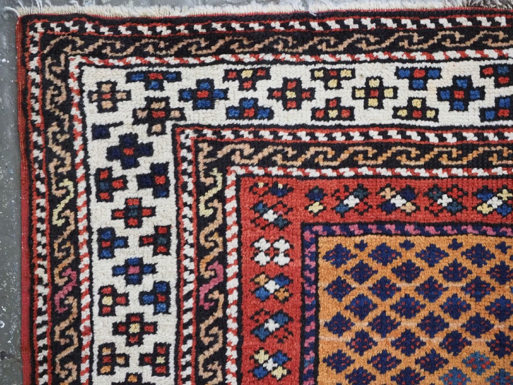A small square antique hand-knotted wool rug featuring a mustard yellow lattice central field surrounded by red and ivory geometric borders. View V