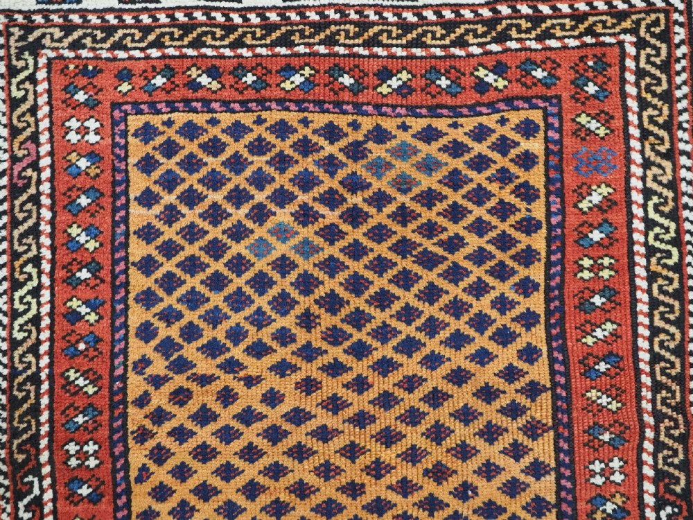A small square antique hand-knotted wool rug featuring a mustard yellow lattice central field surrounded by red and ivory geometric borders. View VI