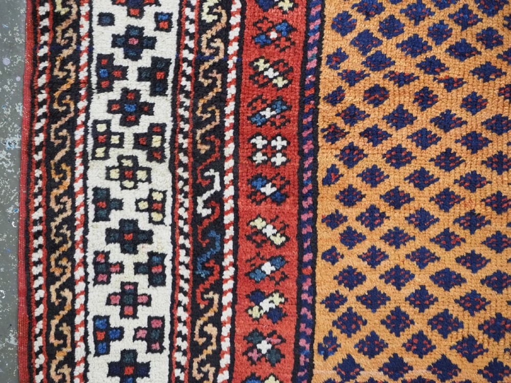 A small square antique hand-knotted wool rug featuring a mustard yellow lattice central field surrounded by red and ivory geometric borders. View VII