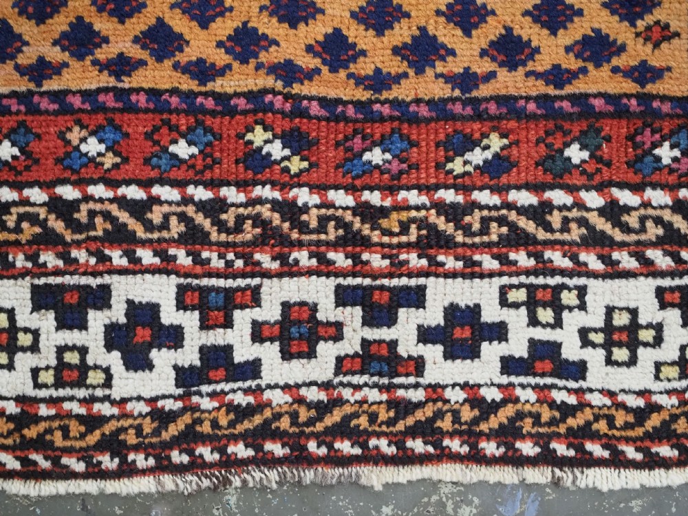 A small square antique hand-knotted wool rug featuring a mustard yellow lattice central field surrounded by red and ivory geometric borders. View IX
