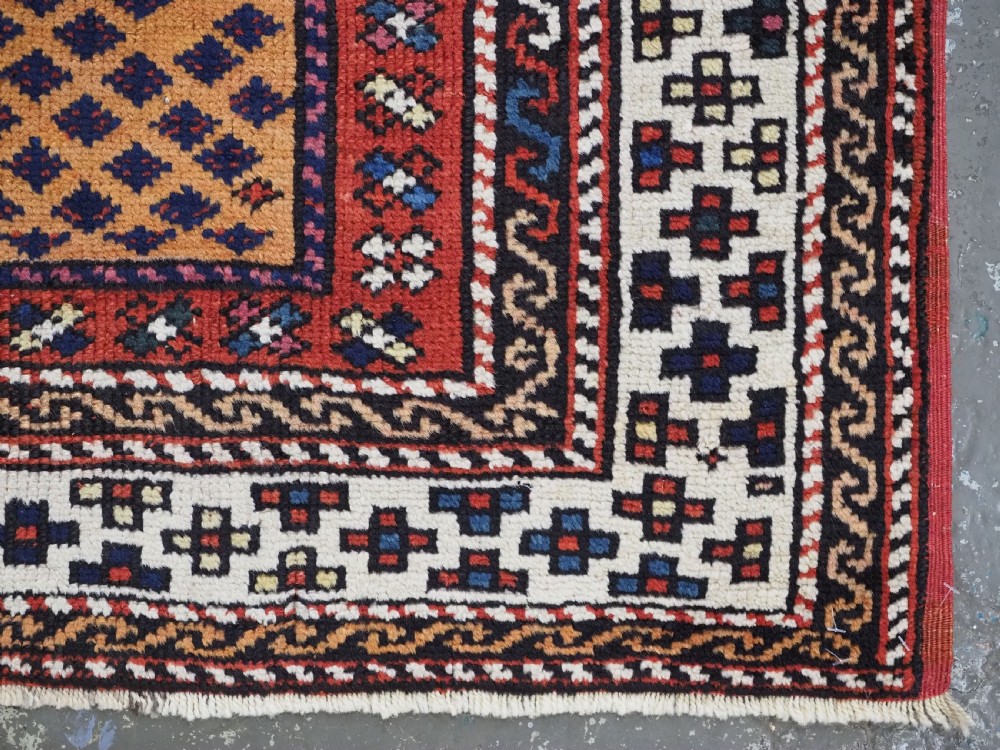 A small square antique hand-knotted wool rug featuring a mustard yellow lattice central field surrounded by red and ivory geometric borders. View X