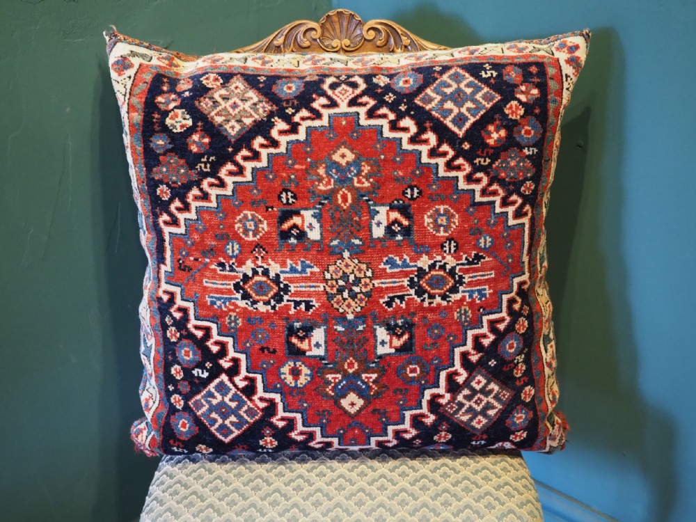 A square handwoven wool carpet cushion featuring a central geometric diamond motif in terracotta red, navy blue, and cream tribal patterns. View II