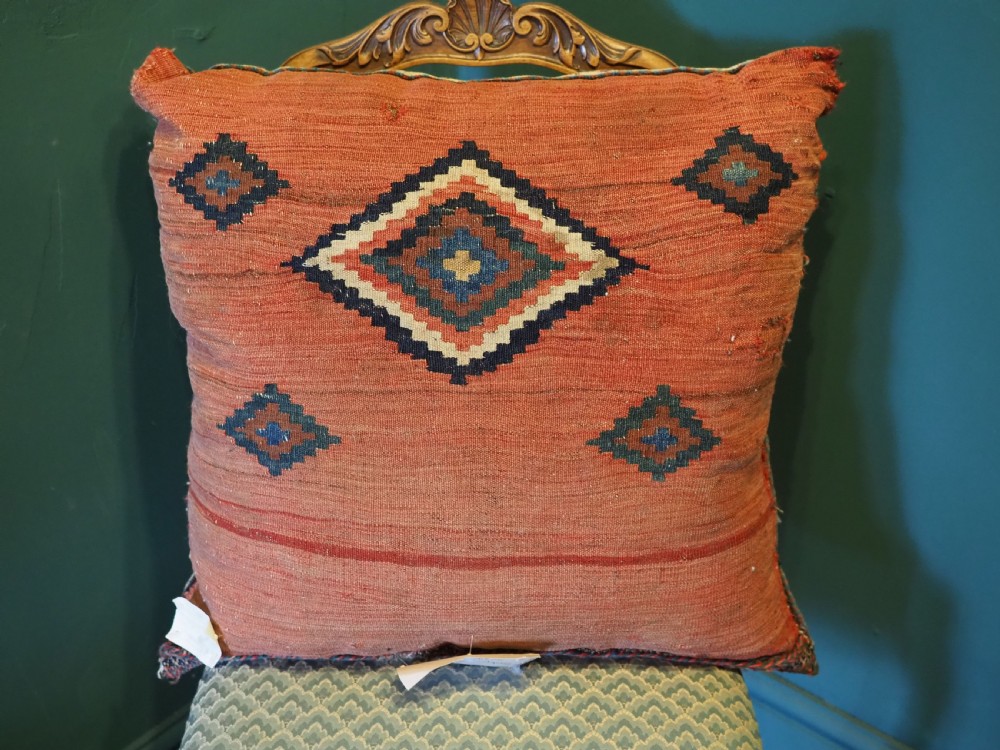 A square handwoven wool carpet cushion featuring a central geometric diamond motif in terracotta red, navy blue, and cream tribal patterns. View III