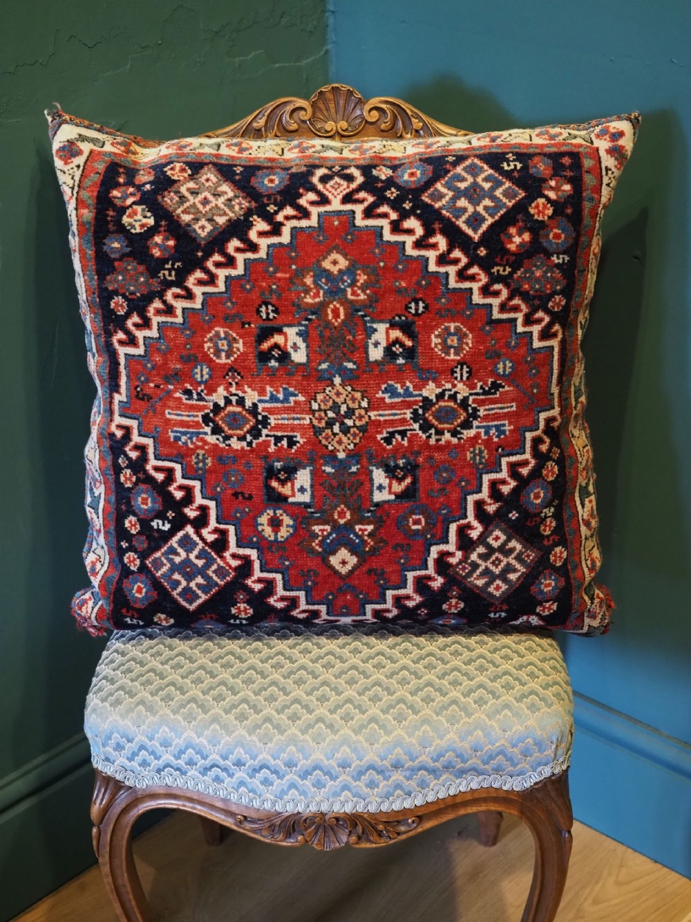 A square handwoven wool carpet cushion featuring a central geometric diamond motif in terracotta red, navy blue, and cream tribal patterns.
