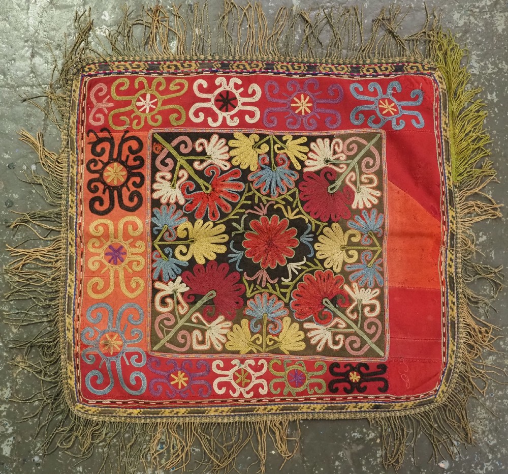 A square antique Uzbek Lakai tribe ilgich embroidery featuring multi-coloured floral and scroll motifs on a red background with fringed edges.