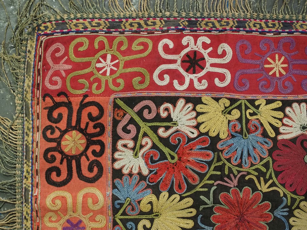 A square antique Uzbek Lakai tribe ilgich embroidery featuring multi-coloured floral and scroll motifs on a red background with fringed edges. View III