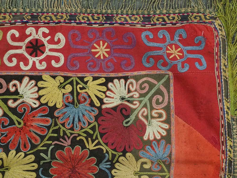 A square antique Uzbek Lakai tribe ilgich embroidery featuring multi-coloured floral and scroll motifs on a red background with fringed edges. View V