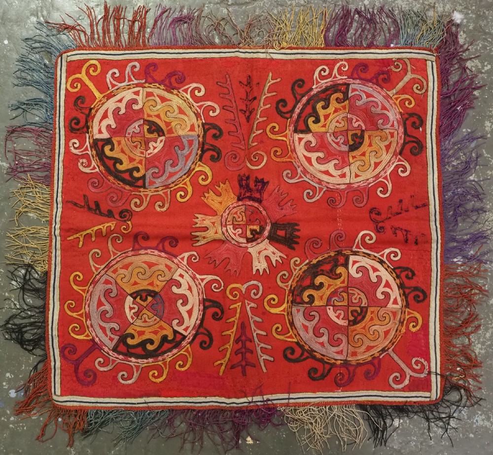 A square red textile wall hanging adorned with hand-embroidered circular solar medallions, spiral motifs, and multi-coloured fringe borders.