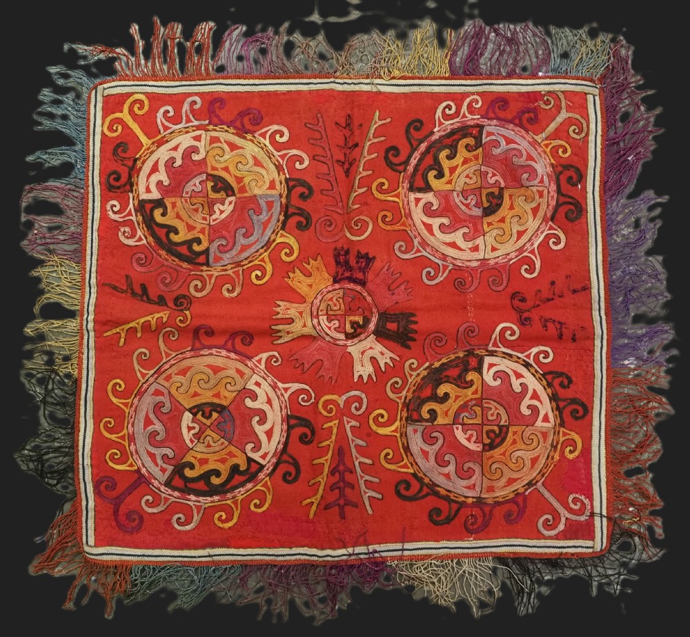 A square red textile wall hanging adorned with hand-embroidered circular solar medallions, spiral motifs, and multi-coloured fringe borders. View II