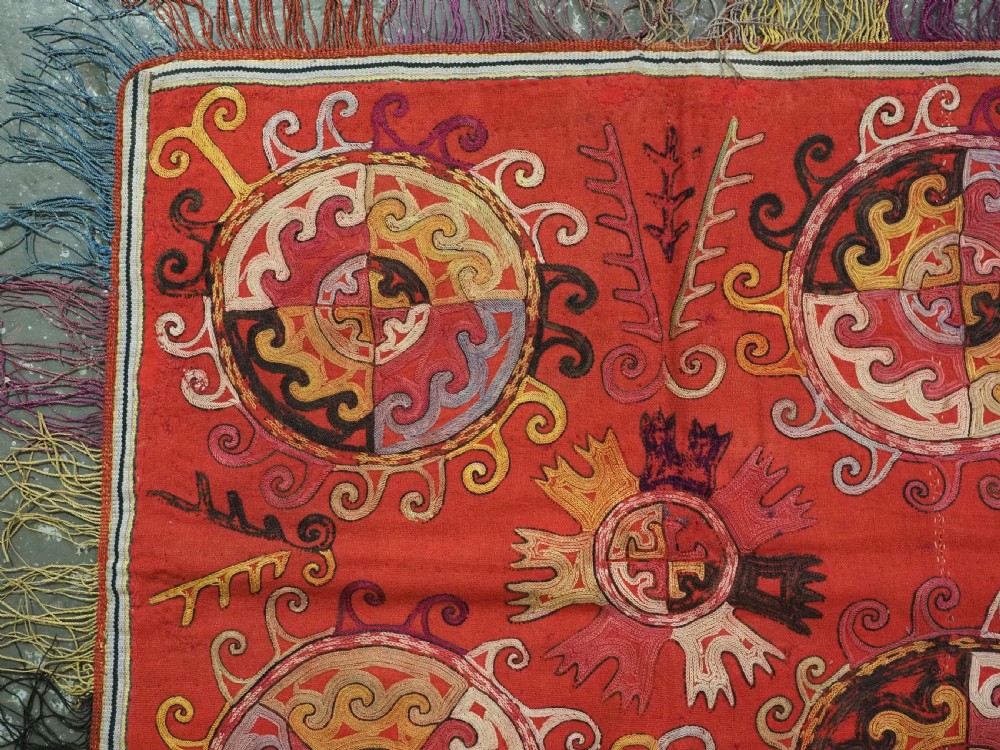 A square red textile wall hanging adorned with hand-embroidered circular solar medallions, spiral motifs, and multi-coloured fringe borders. View III