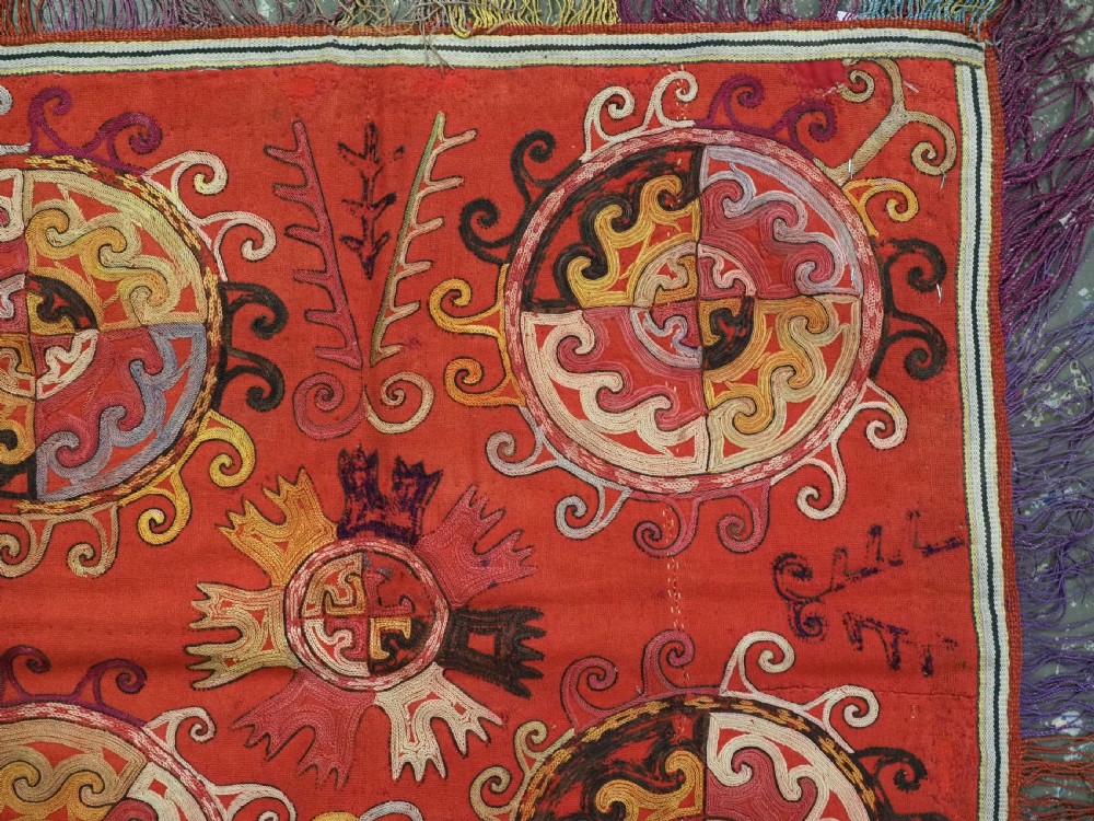 A square red textile wall hanging adorned with hand-embroidered circular solar medallions, spiral motifs, and multi-coloured fringe borders. View IV