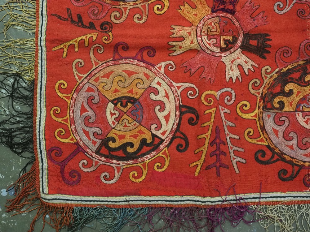 A square red textile wall hanging adorned with hand-embroidered circular solar medallions, spiral motifs, and multi-coloured fringe borders. View V
