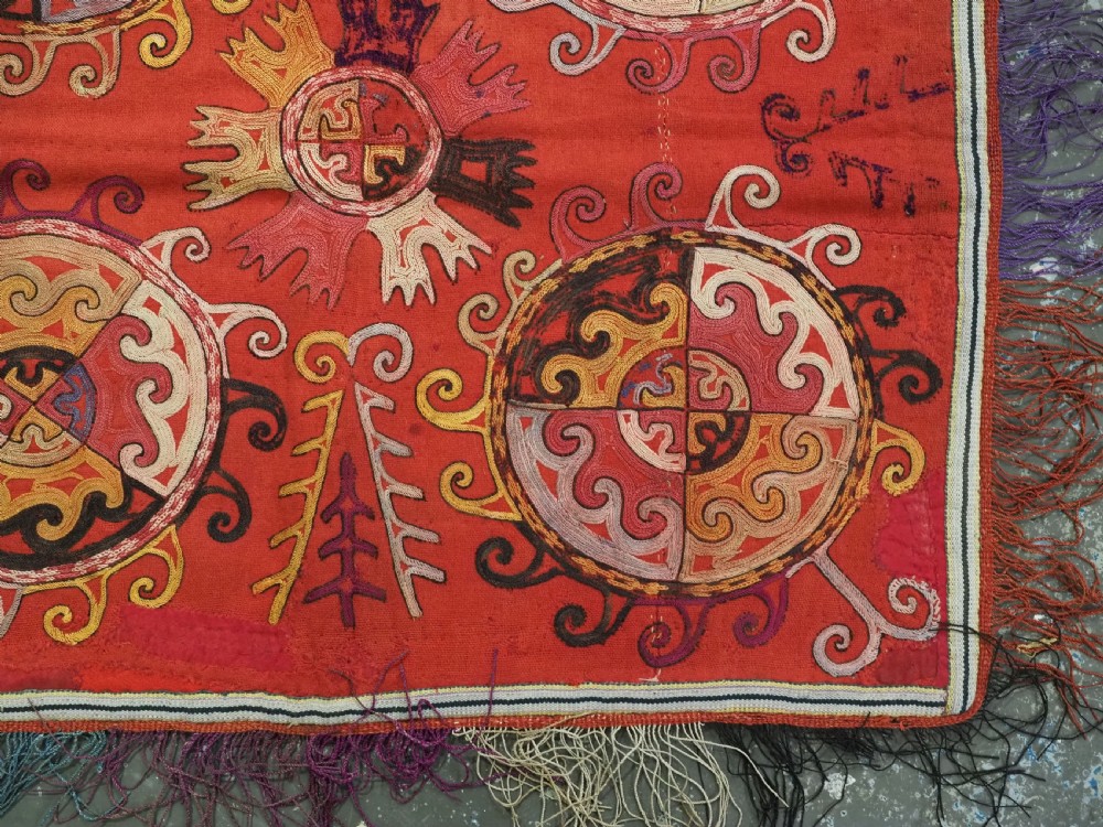 A square red textile wall hanging adorned with hand-embroidered circular solar medallions, spiral motifs, and multi-coloured fringe borders. View VI