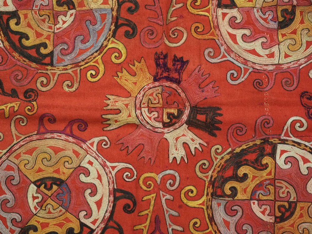 A square red textile wall hanging adorned with hand-embroidered circular solar medallions, spiral motifs, and multi-coloured fringe borders. View VII