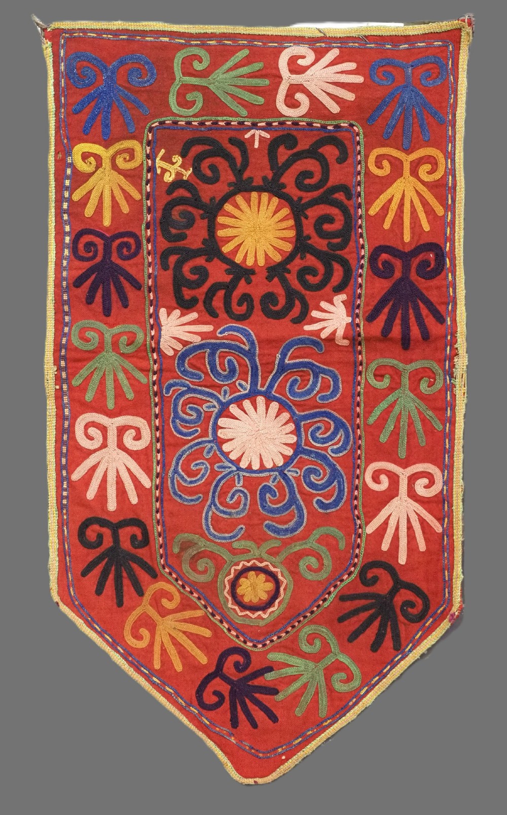 A rectangular red embroidered textile panel with a pointed lower rim, decorated with multi-coloured swirling horn motifs and central rosettes in blue, green, pink, and yellow thread.