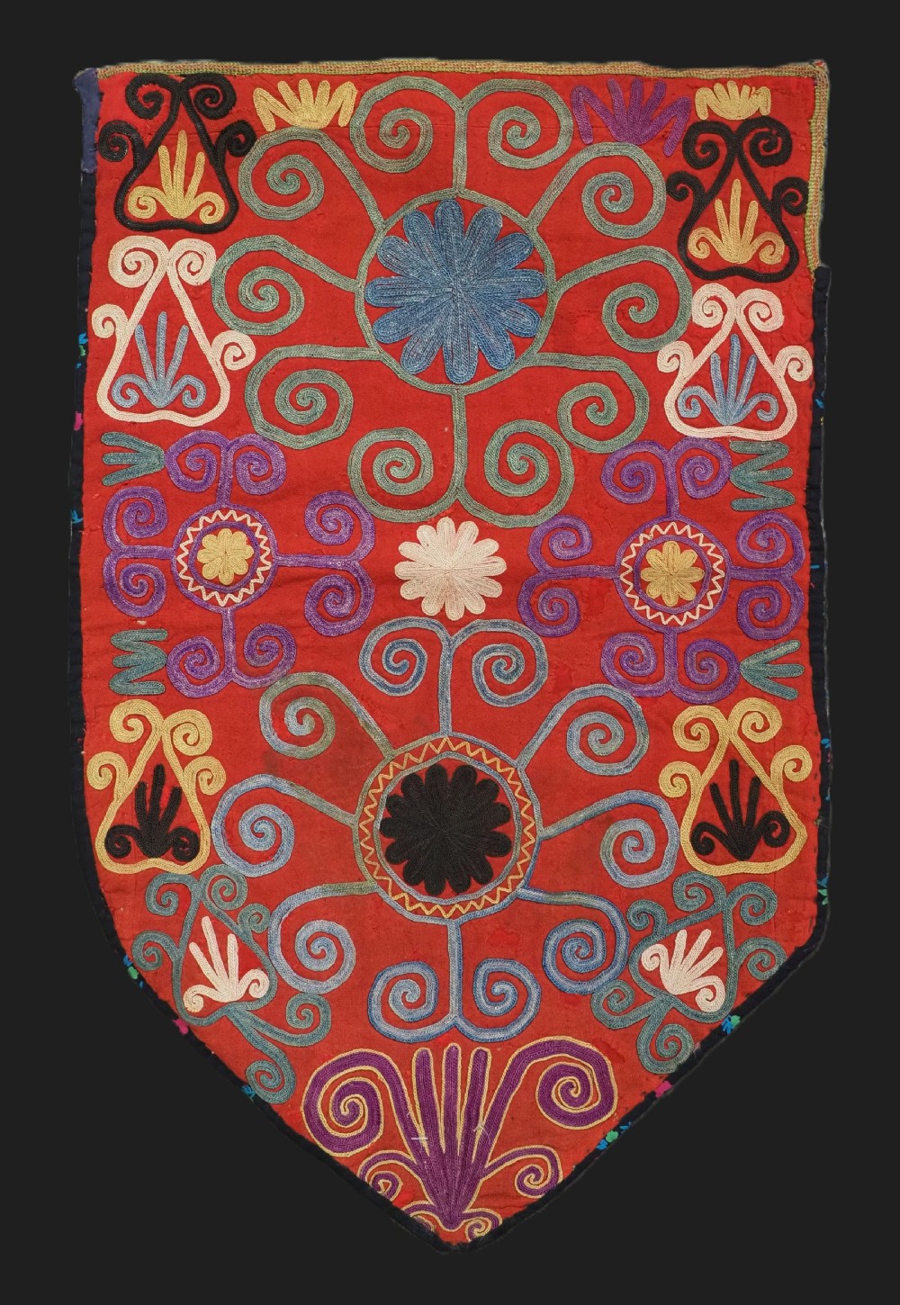 A shield-shaped red Central Asian textile featuring dense silk embroidery with swirling scroll motifs and floral rosettes.