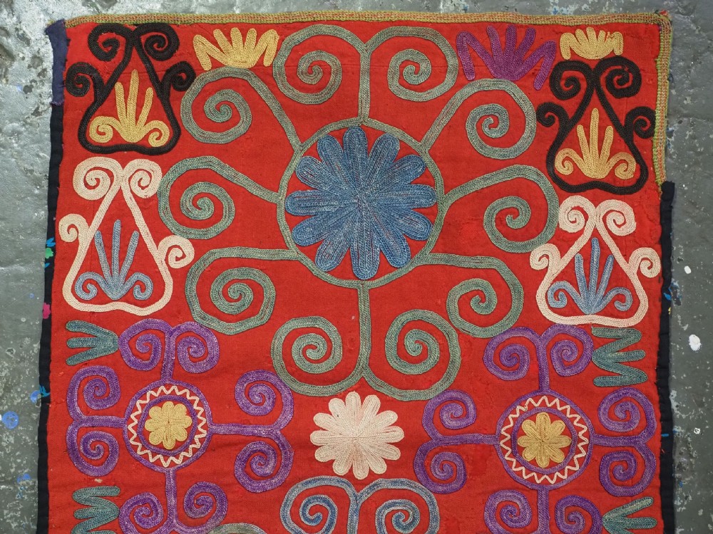 A shield-shaped red Central Asian textile featuring dense silk embroidery with swirling scroll motifs and floral rosettes. View II