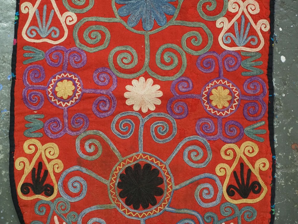 A shield-shaped red Central Asian textile featuring dense silk embroidery with swirling scroll motifs and floral rosettes. View III