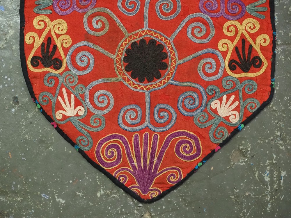 A shield-shaped red Central Asian textile featuring dense silk embroidery with swirling scroll motifs and floral rosettes. View IV