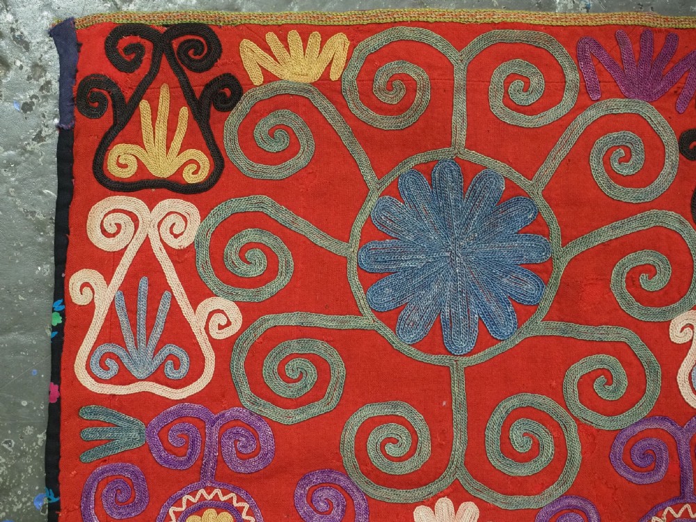 A shield-shaped red Central Asian textile featuring dense silk embroidery with swirling scroll motifs and floral rosettes. View V